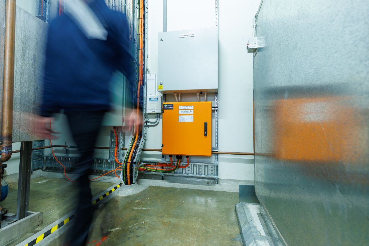 A blurred technician walking past wall-mounted electrical control panels and cabling in an industrial plant room.