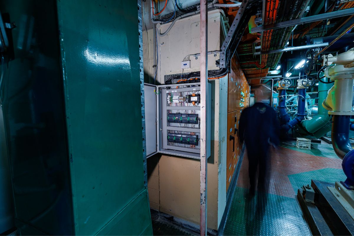 An open electrical control panel inside a mechanical plant room, with pipes, cabling and a technician moving through the space.
